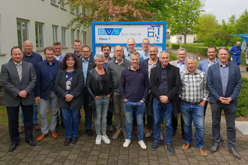 Eine Gruppe von Männern und Frauen steht vor dem Gebäude der SLV Halle mit einem DVS-Schild. Sie posieren für ein Gruppenfoto auf dem gepflasterten Vorplatz bei sonnigem Wetter.