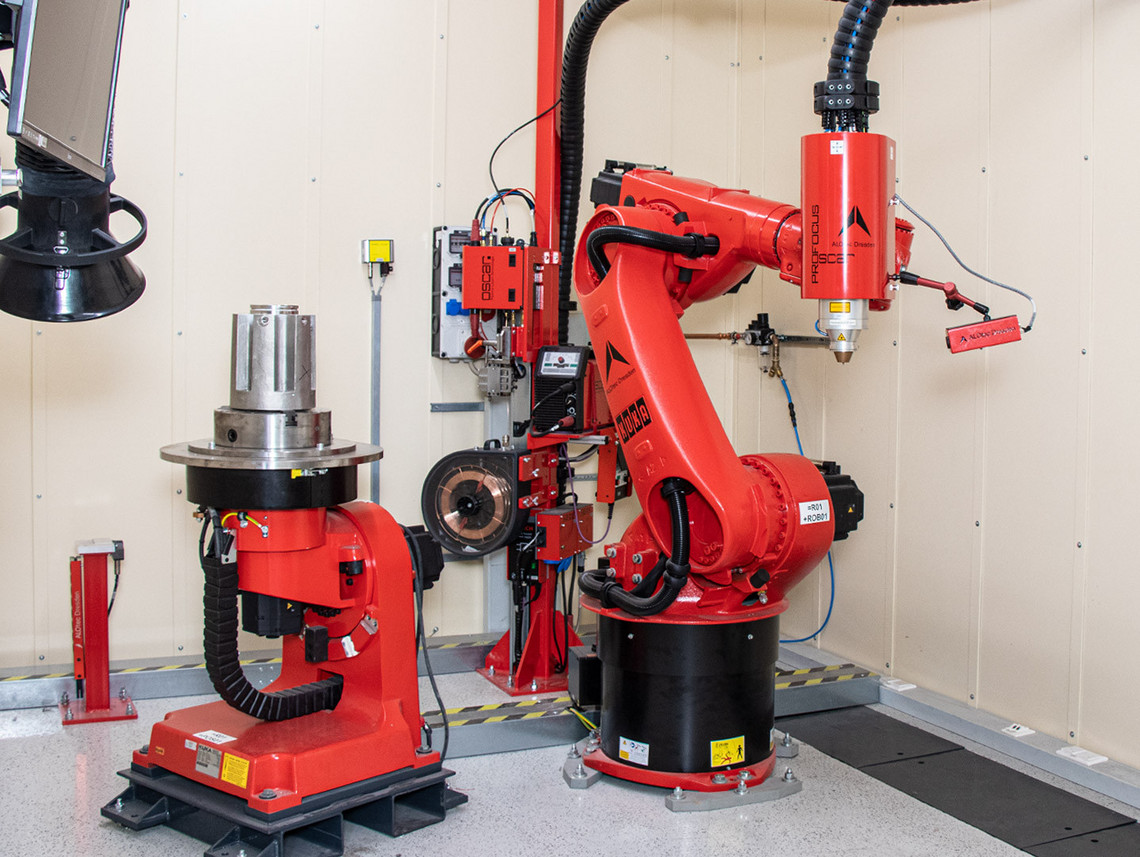 Ein roter Industrieroboter mit Additive-Manufacturing-Toolkopf steht in einer automatisierten Zelle. Daneben befindet sich ein Werkstückhalter mit eingespanntem Metallbauteil.
