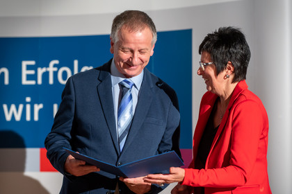 Ein Mann in Anzug mit blauer Krawatte und eine Frau in rotem Blazer lächeln sich an, während sie gemeinsam eine geöffnete Urkunde bei einer Auszeichnungsveranstaltung betrachten.