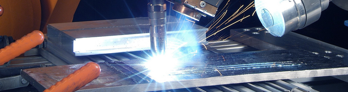 Ein orangefarbener Industrieroboter führt mit einem komplexen Schweißkopf automatisiert einen Lichtbogenschweißprozess durch. Funken fliegen, das Werkstück ist auf einem metallischen Positionierer fixiert, Kabel und Sensoren sind angeschlossen.