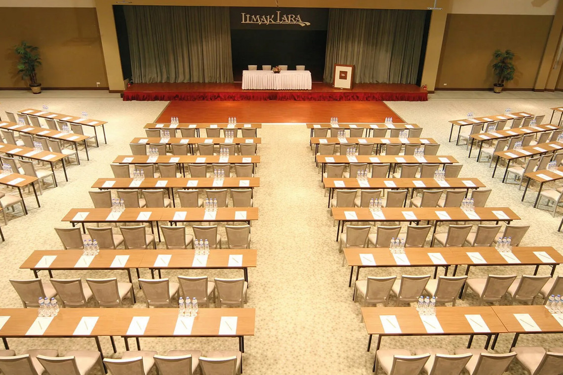 Konferenzsaal mit vorbereiteten Tischen, Wasserflaschen und Schreibmaterialien, Bühne mit Rednerpult im Hintergrund
