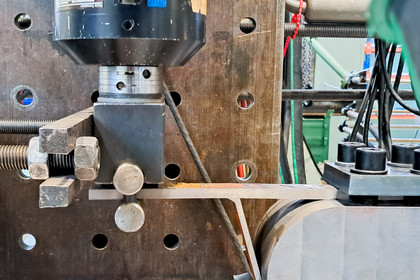 Detailaufnahme eines mechanischen Prüfstands mit eingespanntem Metallprüfkörper bei einem Biegeversuch. Schraubverbindungen, Sensoren und Führungselemente sind sichtbar.