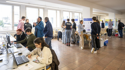 Mehrere Personen befinden sich in einem hellen Raum mit großen Fenstern, diskutieren vor Posterwänden oder arbeiten an Laptops; vorne links sitzen zwei Frauen und ein Mann an Tischen, im Hintergrund stehen kleinere Gruppen im Gespräch