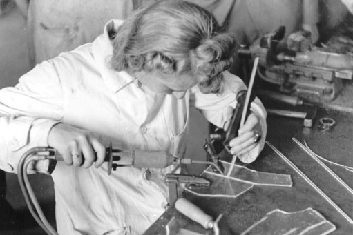 Schwarz-weiß-Fotografie einer Frau in Arbeitskittel beim Schweißen von Kunststoffteilen an einem Werktisch, konzentriert führt sie eine Flamme und ein Werkzeug an ein eingespanntes Werkstück.