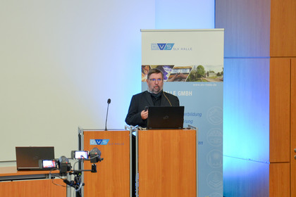 Mann spricht auf einer Bühne hinter einem Rednerpult mit SLV-Halle-Logo, vor ihm Laptop und Mikrofon, im Hintergrund Roll-Up der SLV Halle und 