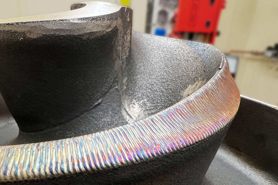 Nahaufnahme einer metallischen Oberfläche mit gleichmäßiger Schweißraupe, die sich spiralförmig um ein rotationssymmetrisches Bauteil legt. Die Oberfläche zeigt irisierende Farbtöne.