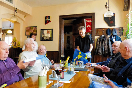 Mehrere ältere Männer sitzen und stehen in einem Gemeinschaftsraum, Steffen Wagner überreicht ein Buch an eine andere Person, weitere Personen beobachten die Szene, im Hintergrund Türen, Garderobe und Dekorationen 