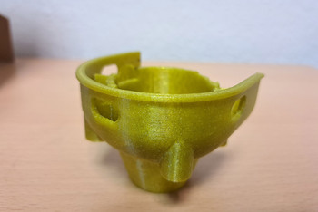 Gelbes 3D-gedrucktes Kunststoffmodell eines Bauteils mit mehreren Öffnungen und Verstärkungsrippen steht auf einer Holzoberfläche; es stellt eine rekonstruierte Version eines beschädigten Gussstücks dar.