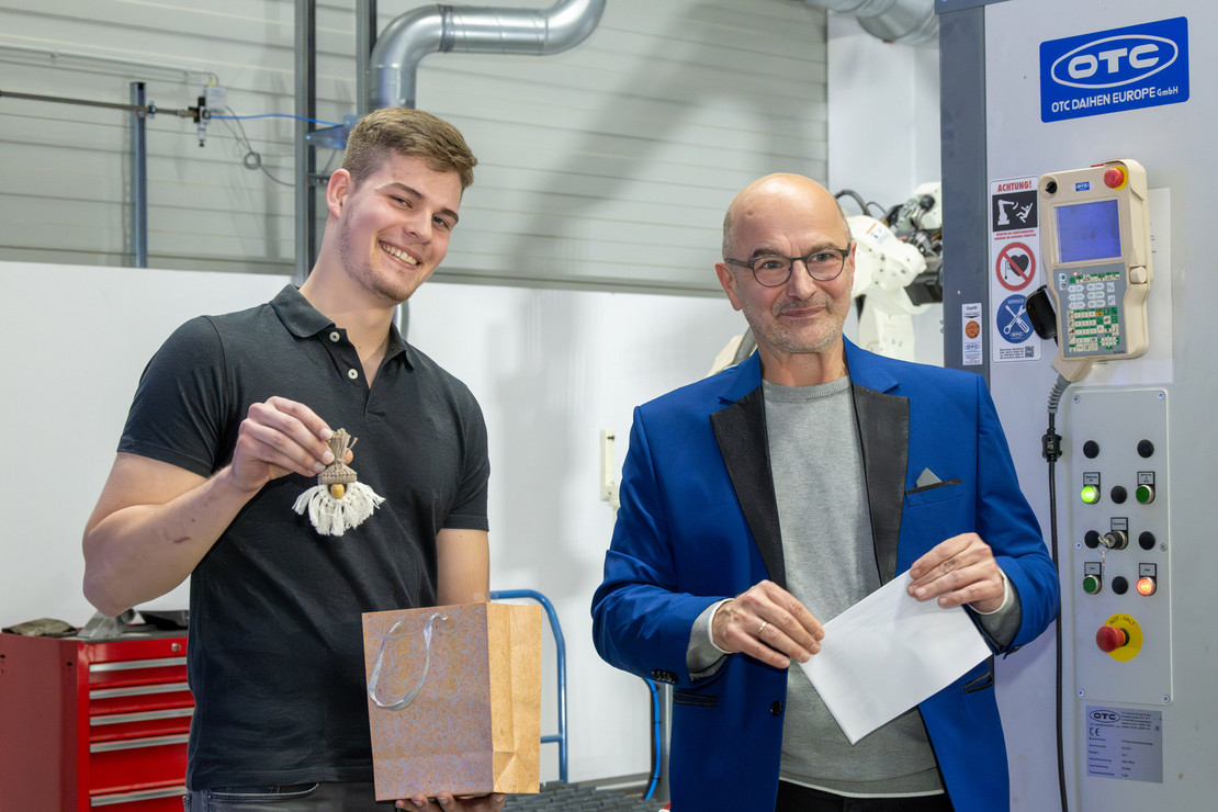 Ein junger Mann mit Geschenktüte und kleinem Schutzengel steht neben einem älteren Mann im blauen Sakko mit Umschlag in der Hand; beide lächeln, im Hintergrund sind industrielle Maschinen und Lüftungsrohre zu sehen.