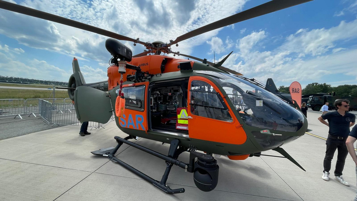 Such- und Rettungshubschrauber mit orange-grüner Lackierung und offener Seitentür steht auf dem Flugfeld, Innenausstattung sichtbar, Personen daneben