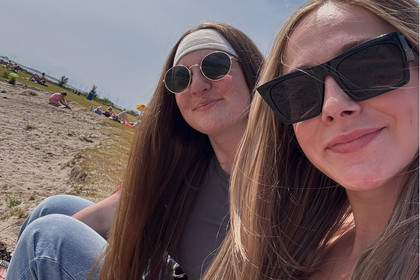 Zwei junge Frauen mit langen Haaren und Sonnenbrillen sitzen entspannt am See, im Hintergrund eine sandige Böschung
