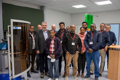 Gruppenfoto in einem technischen Schulungsraum, viele tragen Namensschilder, links im Bild ein Gerät mit Schutzschild und SLV-Logo, im Hintergrund grüne Vorhänge und moderne Einrichtung.