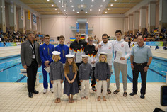 Gruppe  steht am Rand eines Schwimmbeckens, darunter sechs Sportler (alle Männer) in Nationalkleidung, zwei Offizielle und vier Kinder in traditioneller Kleidung mit Matrosenmützen, im Hintergrund Zuschauertribünen und ein Sprungturm.