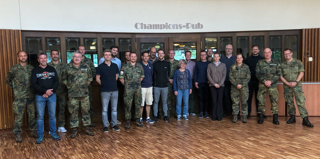 Gruppenfoto mit 23 Personen, teils in ziviler Kleidung, teils in Bundeswehr-Uniform, aufgenommen vor dem Eingang zum „Champions-Pub“ mit Holzfußboden und Glasfront im Hintergrund.