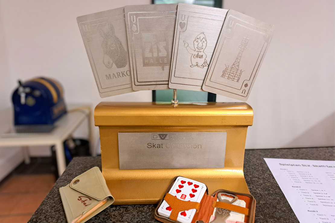 Nahaufnahme einer Holzbox mit eingesetzten Spielkarten und Pokalen, daneben orangefarbene Kartenpackungen.