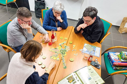 Vier Personen sitzen an einem Tisch und spielen ein Brettspiel, konzentrierter Blick auf Spielfeld und Karten.