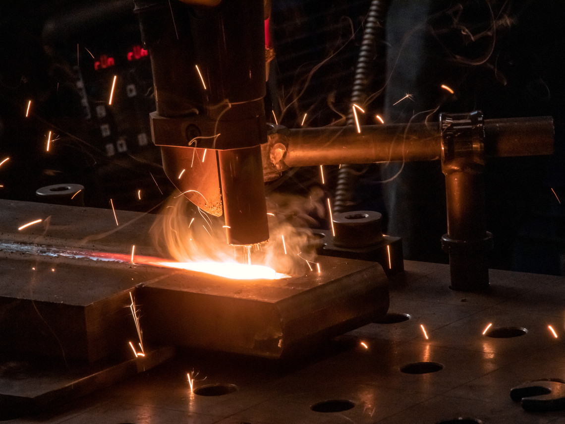 Automatisiertes Schweißgerät erzeugt eine helle Schweißnaht auf einer dicken Metallplatte; Funken fliegen in alle Richtungen, im Hintergrund sind Schläuche und Bedienelemente sichtbar.