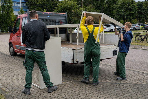Drei männliche Personen verladen weiße Möbelteile auf die Ladefläche eines Transporters. Zwei tragen gemeinsam ein Tischgestell, die dritte Person steht neben einem hohen Schrankelement; alle tragen grüne Arbeitshosen oder Latzhosen, teilweise mit Arbeitshandschuhen.