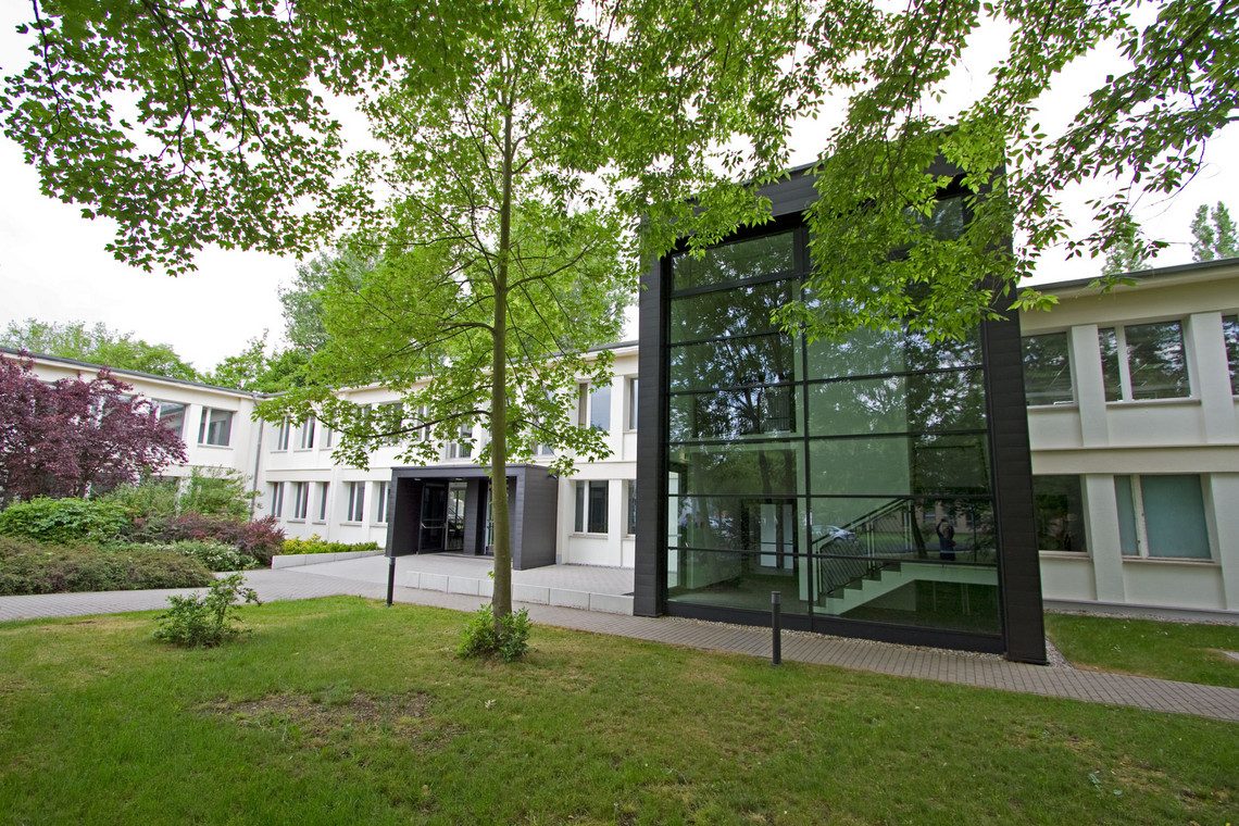 Ein modernes Verwaltungs- oder Institutsgebäude mit weißer Fassade und einem auffälligen, schwarz umrahmten Glaseingang steht hinter gepflegtem Grün. Ein Baum in der Bildmitte spendet Schatten, ein gepflasterter Weg führt zum Haupteingang.