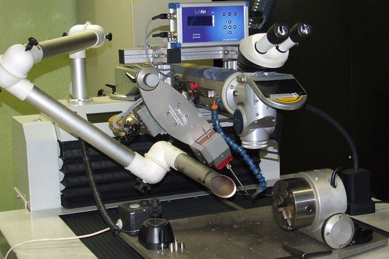 Ein automatisiertes Laserschweißsystem mit integriertem Mikroskop und flexiblem Roboterarm ist auf einem Arbeitstisch aufgebaut. Das Werkstück ist in einer Drehvorrichtung eingespannt, ein Display zur Steuerung befindet sich im Hintergrund.