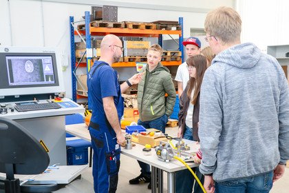 Drei Jungen und ein Mädchen stehen an einem Arbeitstisch mit elektronischen Bauteilen und Werkzeugen in einer Werkhalle. Ein Mann in Arbeitskleidung erklärt den Aufbau, während die anderen Personen den Tisch betrachten.