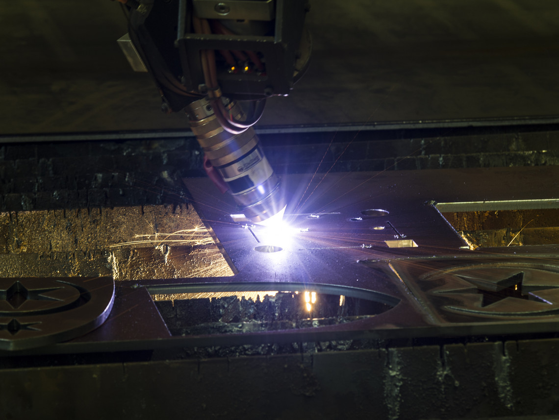Detailaufnahme eines Plasmaschneidkopfs, der mit hellem Lichtstrahl präzise Formen aus einer Metallplatte schneidet; rund ausgesparte Sternsymbole sind bereits sichtbar.
