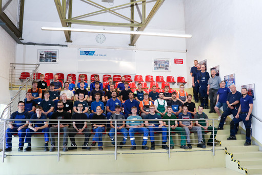Gruppenfoto mit etwa 60 Auszubildenden und Ausbildenden auf Tribünenstufen im SLV-Ausbildungszentrum; überwiegend Männer, einige Frauen, alle in Arbeitskleidung, im Hintergrund rote Sitzschalen und das Logo der SLV Halle.