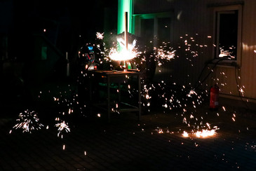 Bei einer Vorführung im Außenbereich sprühen zahlreiche Funken von einem beleuchteten Metallbearbeitungsplatz in die Dunkelheit.