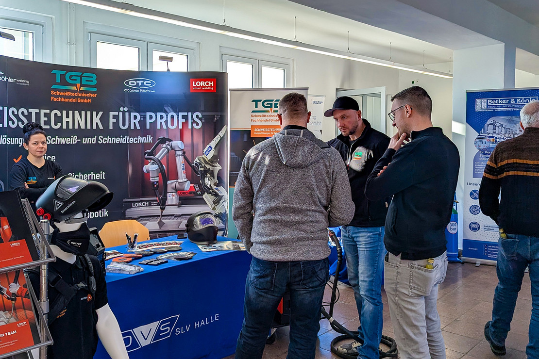 Mehrere Personen stehen in einem Ausstellungsbereich an einem Informationsstand der Schweißtechnik, auf dem Tisch liegen Informationsmaterialien, im Hintergrund sind Rollups und eine Präsentationswand mit technischen Motiven zu sehen
