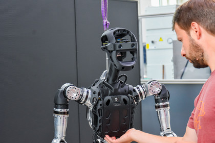 Ein Mann in rotem T-Shirt hält seine Hand unter den Oberkörper eines humanoiden Roboters mit schwarzen Gehäuseteilen und metallischen Armen, der an einem violetten Hebegurt hängt.
