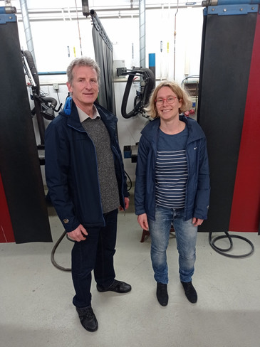 Ein Mann und eine Frau stehen lächelnd nebeneinander in einer Werkstatt, beide tragen blaue Jacken, im Hintergrund sind technische Vorrichtungen sichtbar.