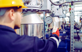 Arbeiter mit gelbem Schutzhelm und Handschuh überprüft ein Manometer an einer großen metallischen Industrieanlage.
