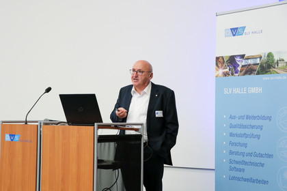 Ein Mann im Anzug spricht vor einem Publikum bei einer Fachveranstaltung; links im Bild befinden sich Titel und Logos der Veranstaltung