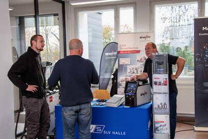 Drei Männer unterhalten sich an einem Infostand mit SLV-Halle-Tischdecke über Schweißtechnik, auf dem Tisch steht ein EWM-Schweißgerät; im Hintergrund sind Roll-ups und Prospektständer platziert.