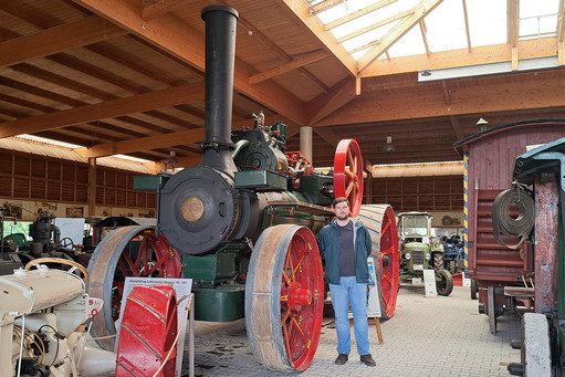 Eine historische Maschine mit großen Speichenrädern, grünem Kessel und hohem schwarzem Schornstein steht in einer offenen Halle mit Holzdach.