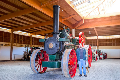 Eine historische Maschine mit großen Speichenrädern, grünem Kessel und hohem schwarzem Schornstein steht in einer offenen Halle mit Holzdach.