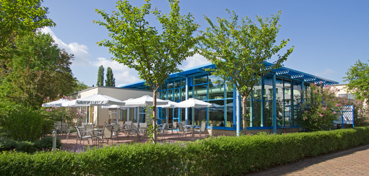 Außenansicht eines verglasten Veranstaltungsbereichs mit blauen Stahlträgern. Davor eine Terrasse mit mehreren Sitzgruppen und weißen Sonnenschirmen, umgeben von blühenden Sträuchern und jungen Bäumen. Im Vordergrund eine gepflegte Hecke und gepflasterter Weg.