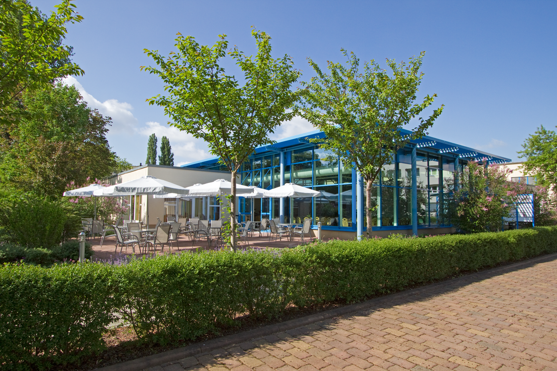Außenansicht eines verglasten Veranstaltungsbereichs mit blauen Stahlträgern. Davor eine Terrasse mit mehreren Sitzgruppen und weißen Sonnenschirmen, umgeben von blühenden Sträuchern und jungen Bäumen. Im Vordergrund eine gepflegte Hecke und gepflasterter Weg.