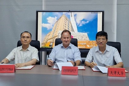 Drei Männer sitzen an einem Konferenztisch vor Namensschildern mit chinesischen Schriftzeichen und unterzeichnen Dokumente, im Hintergrund zeigt ein Bildschirm ein Hochhaus mit der Aufschrift „THE XD“ und den Hinweis auf ein Innovationszentrum.