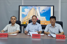 Drei Männer sitzen an einem Konferenztisch vor Namensschildern mit chinesischen Schriftzeichen und unterzeichnen Dokumente, im Hintergrund zeigt ein Bildschirm ein Hochhaus mit der Aufschrift „THE XD“ und den Hinweis auf ein Innovationszentrum.