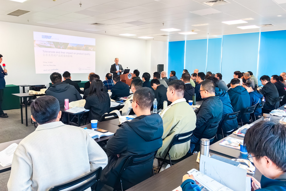 Großer Seminarraum mit zahlreichen Teilnehmenden, die an Tischen sitzen und einer Präsentation mit Folien und Vortrag folgen.