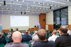 Ein gut besuchter Vortragssaal mit zahlreichen männlichen Teilnehmenden, die auf eine Bühne mit Redner und groß projizierter Präsentationsfolie blicken; der Raum ist modern ausgestattet mit Lichtpaneelen und großen Fenstern.