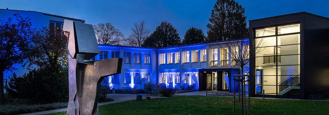 Gebäude der SLV Halle GmbH bei Nacht, in blauer Beleuchtung, mit Skulptur im Vordergrund und beleuchtetem Eingangsbereich.