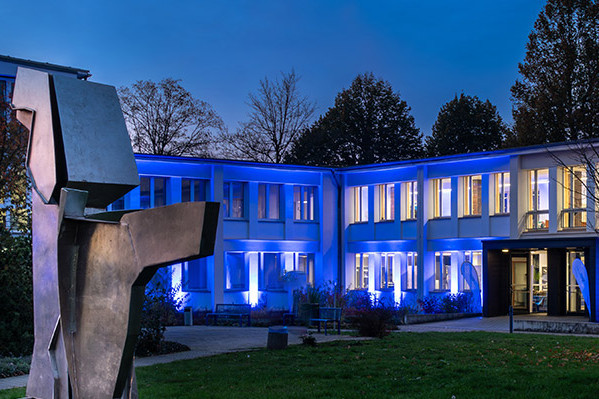 Gebäude der SLV Halle GmbH bei Nacht, in blauer Beleuchtung, mit Skulptur im Vordergrund und beleuchtetem Eingangsbereich.