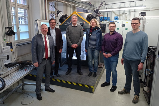 Sechs Personen, fünf Männer und eine Frau, stehen in einem Laborraum vor einem Prüfgerät mit schwarzer Umhausung, Kabeln und Steuertechnik im Hintergrund