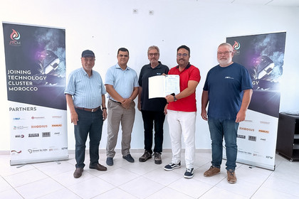 Fünf Männer stehen vor zwei Roll-up-Bannern des Joining Technology Cluster Morocco in einem hellen Raum, der Mann in der Mitte hält ein aufgeschlagenes Zertifikat in die Kamera.