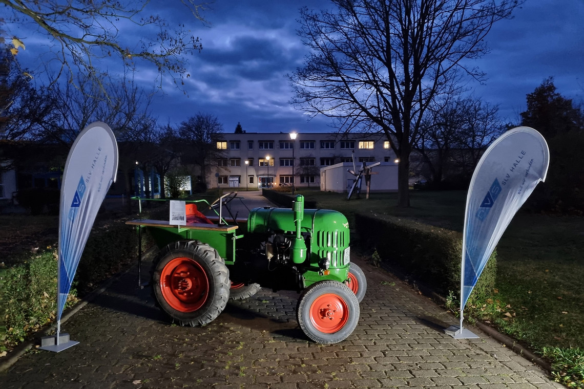 Ein grüner Oldtimer Lanz-Traktor mit roten Felgen steht bei Dunkelheit beleuchtet zwischen zwei DVS SLV Halle-Bannern auf einem gepflasterten Weg; im Hintergrund ist ein beleuchtetes Gebäude zu sehen.