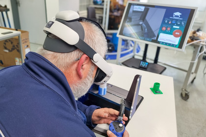 Ein Mann sitzt an einem Elektronikarbeitsplatz und trägt eine VR-Brille, während er mit einem Lötkolben an einem kleinen Bauteil arbeitet. Vor ihm stehen Monitore mit technischen Anzeigen, auf dem Tisch liegen Werkzeuge, Kabel und elektronische Komponenten.
