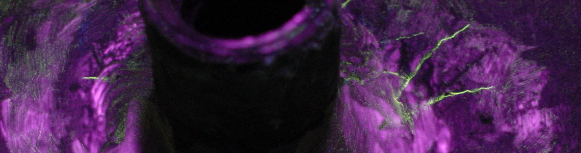 Magnetpulverprüfung bei UV-Licht zeigt feine Rissanzeigen in grün entlang einer Schweißverbindung um ein zentrales Rohr, umgeben von fluoreszierendem Prüfpulver.