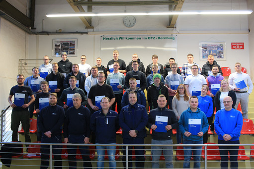 Gruppenfoto von etwa 35 Personen auf Tribüne, viele halten Zertifikate in der Hand, Banner mit Schriftzug „Herzlich Willkommen im BTZ Bernburg“ im Hintergrund